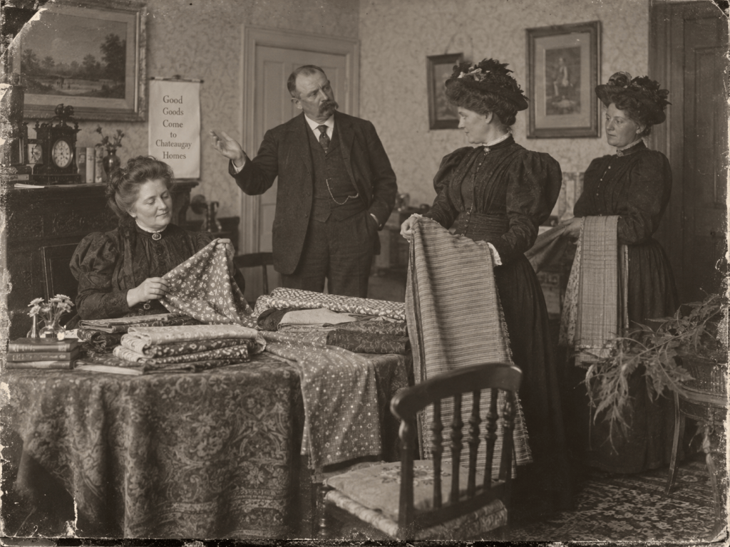 A vintage setting featuring three women and a man engaged in a discussion about fabrics. The women are examining different patterned cloths on a table, while the man gestures as he speaks. The backdrop includes decorative wallpaper and framed artwork.