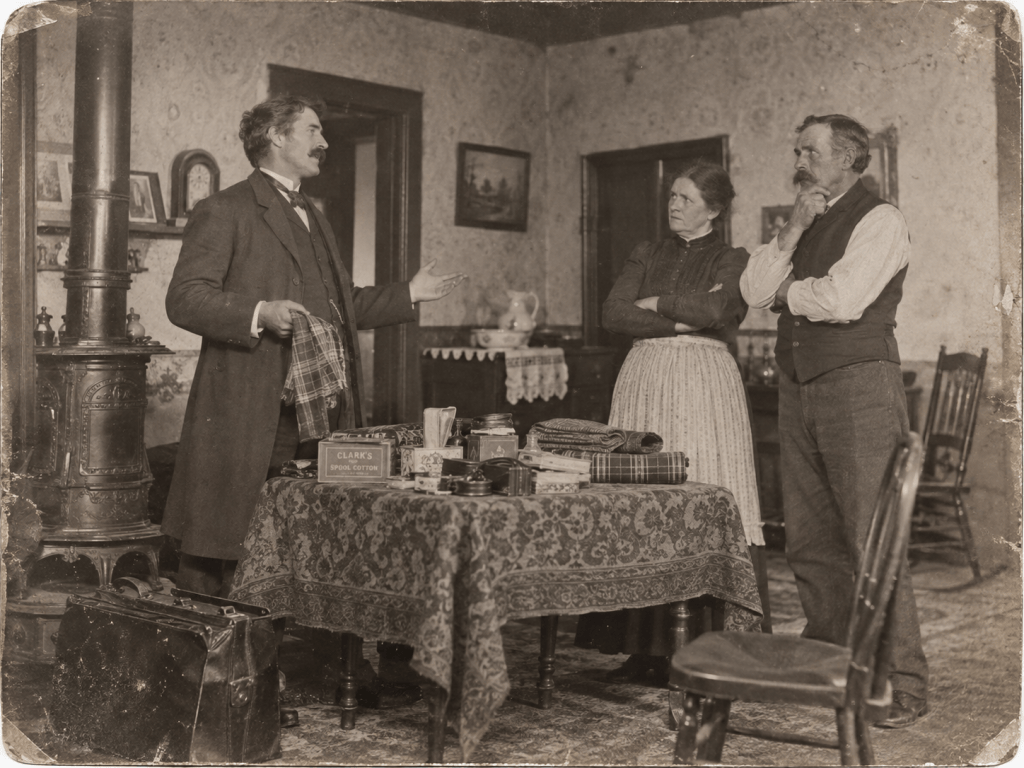 A man in a coat gestures while holding a cloth, talking to a woman and a man who appear skeptical, inside a vintage-style room with antique furnishings and a wood stove.